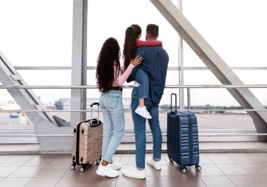 Aile gezisi. Küçük kızıyla birlikte havaalanının penceresinde duran anne, baba ve kız çocuğunun birlikte uçağa binmeyi beklerken manzaranın tadını çıkardıkları arka manzara,