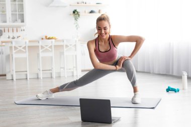 İnternette sabah antrenmanı, ders, covid-19 salgınında kendini soyutlama. Oturma odasının iç kısımlarında gülümseyen sarışın bayan paspasın üzerinde bacaklar için egzersiz yapıyor, dizüstü bilgisayara bakıyor.