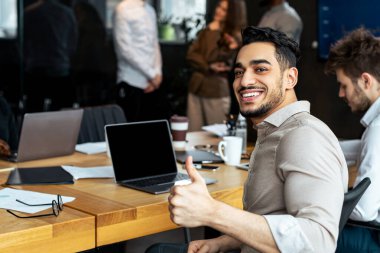 Başarılı Buluşma Konsepti. Modern ofiste meslektaşlarıyla birlikte masasında oturan, işaret işareti yapan, poz veren ve kameraya bakan kendinden emin genç iş adamının portresi.