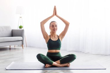 Meditasyon, nefes alma egzersizleri, dinlenme. Sağlıklı yaşam tarzı ve sağlık konsepti. Sakin, sarışın, spor kıyafetli bir kadın. Lotus pozisyonunda. Sabahları oturma odasında yoga yapıyor.