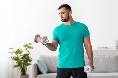 Mavi tişörtlü beyaz erkek sporcu dambılları kaldırıyor ve minimalist oturma odasının iç kısımlarında pazılar için egzersiz yapıyor. Evde tek başına eğitim, covid-19 sırasında vücut bakımı ve spor