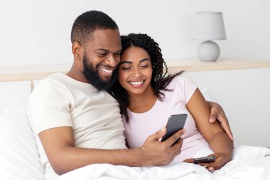 Smiling happy young african american boyfriend and girlfriend hugging, sitting on bed in bedroom interior, surfing in internet at phone. Video call, new normal, communication remotely, great offer