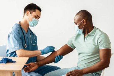 Tıp merkezine enjeksiyon, aşı kampanyası. Koruyucu maskeli ve üniformalı bin yıllık doktor, klinikteki Afro-Amerikan hastasına ilaç ve şırınga ampulleri olan bir masa verdi.