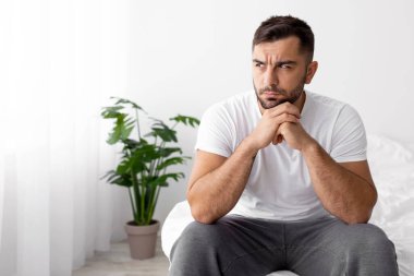 Çaresiz, yalnız, Avrupalı kaslı, kaslı bir adam beyaz yatakta oturur ve yatak odasının içini düşünür. Boşluğa bakar. Stres, sorun, depresyon ve covid-19 salgını sırasında evde kötü haberler