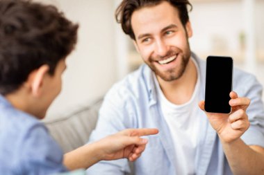 Modern teknoloji konsepti. Babası oğluna siyah ekranlı yeni bir akıllı telefon gösteriyor. Model için yeri olan bir telefon, kopyalama alanı. Çocuk telefonu parmağıyla gösterip koltukta oturuyor.