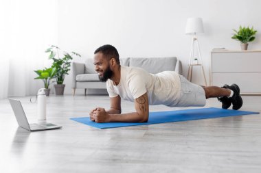 Genç siyah sakallı adamın odanın içinde döşeme döşemesi ve bir şişe su ile dizüstü bilgisayarda antrenman yapmasına sevindim. Çevrimiçi sınıf, koçtan ders, tecrit sırasında eğitim ve vücut bakımı, profil