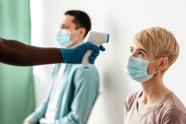 Coronavirus bağışıklığı, tıbbi nüfus aşılama kampanyası. Koruyucu eldivenli Afrikalı Amerikalı erkek hemşire termometre ile klinikte maskeli yetişkin kadın termometreyi ölçüyor.