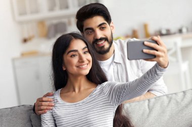Yeni normal, selfie, sosyal uzaklık sırasında görüntülü konuşma. Mutlu çekici genç Arap bayan ve sakallı adam akıllı telefonla fotoğraf çekiyor, web kamerasına bakıyor, oturma odasındaki kanepeye bakıyor, boş bir alanda oturuyor.