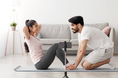 Koçlu dersler, video dersi, karantina sırasında evde eğitim. Gülümseyen bin yıllık Arap adam karına karın egzersizlerinde yardım ediyor. Oturma odasının iç kısmında üç ayaklıya telefonla ateş ediyor.