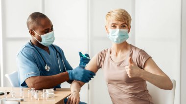 Korunaklı maske takan doktor, koronavirüs enfeksiyonu riski olan kadın hastaların bağışıklığını uyarmak için aşı enjekte ediyor. Milenyum Afrikalı Amerikalı hemşire yetişkin kadınlara iğne yapıyor ve başparmağını gösteriyor.