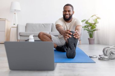 Sakallı, neşeli, Afro-Amerikan bir adam dizüstü bilgisayara bakmak için paspasla bacak esnetiyor. Video sınıfı, spor ekipmanları, spor, spor, sağlık ve vücut bakımı, covid-19 kendi kendini izole etme