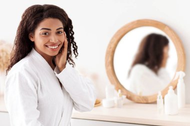 Blogcu, cilt bakımı, rutin prosedür ve spa merkezinden güzel tavsiyeler. Mutlu bin yıllık Afro-Amerikan kadın beyaz önlüklü, yüzü yatak odası, iç kısmı kozmetik ve ayna ile birbirine dokunuyor.