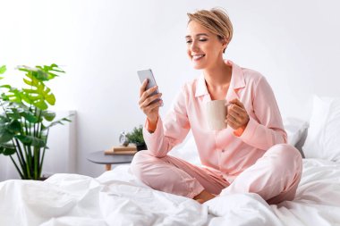 Sabah Vakti. Akıllı telefon kullanan, interneti karıştıran ve elinde bir fincan kahve tutan gülümseyen bir kadın. Pembe ipek gecelikli bayan dinleniyor ve beyaz yatak odasındaki aletlere bakıyor.