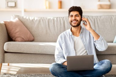 Arap serbest çalışan bir adam dizüstü bilgisayar kullanıyor ve cep telefonuyla konuşuyor, oturma odasında yerde otururken çalışıyor. Her zaman iletişim halinde, uzaktan çalışma ve mobil iletişim, serbest yaşam tarzı