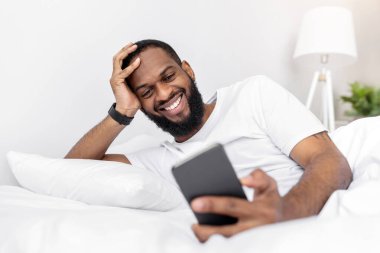 Sakallı neşeli Afrikalı Amerikalı genç adam akıllı telefona bakar, yatak odasının içindeki beyaz yatağa uzanır. Video görüşmesi ve sosyal mesafe, yeni uygulama ve komik video, reklam ve evdeki teklif
