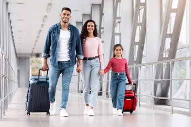 Seyahat sigortası. Kızıyla birlikte havaalanında yürüyen gülümseyen Arap aile, mutlu Orta Doğulu ebeveynler ve el ele tutuşan ve kameraya bakan küçük bayan çocuk,