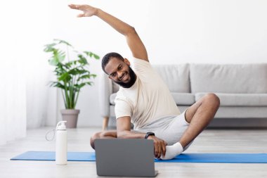 Beyaz giysili mutlu siyah sakallı atlet, paspas üzerinde ellerini esnetiyor ya da egzersiz yapıyor. Oturma odasının iç kısımlarına bakıyor. Koç Covid-19 'la evdeki spor salonunda online ders ve egzersiz.