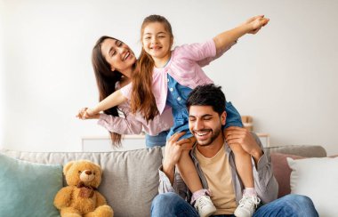 Neşeli, genç, doğulu ebeveynler oturma odasında çocukla eğleniyor, oynuyor, gülüyor ve birlikte vakit geçiriyorlar. Evde kızı olan mutlu bir Arap ailesi.