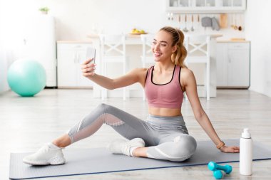 Dostça gülümseyen Kafkas genç sarışın fitness blogcusu telefon kamerasına bakıyor ve sosyal ağlar için canlı yayın yapıyor. Kadın video spor blogu, selfie, yeni normal ve sosyal mesafe