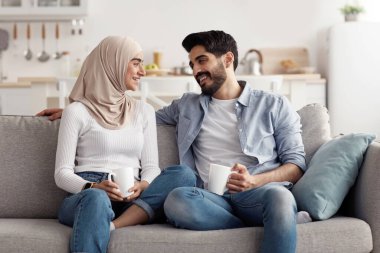 Birlikte eğlenmek, kahvaltıda eğlenmek ve evde boş vakit geçirmek, aile randevusu. Gülümseyen, tesettürlü, tesettürlü, genç, çekici bir Arap kadın ve oturma odasındaki kanepede boş bir alanla konuşan ve bardakları tutan bir adam.