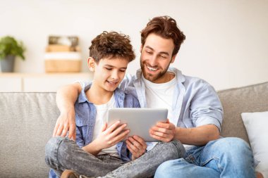 İnternetten alışveriş konsepti. Mutlu baba ve oğlu internete dijital tabletle bakıyorlar, evde birlikte vakit geçiriyorlar, rahat bir koltukta oturuyorlar. Boş aktiviteler, modern teknoloji konsepti