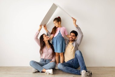 Aile bakımı ve koruma konsepti. Doğulu ebeveynler, yeni evlerinde beyaz duvarın yanında oturan küçük kızlarının üstündeki ellerin sembolik çatısını yapıyorlar.