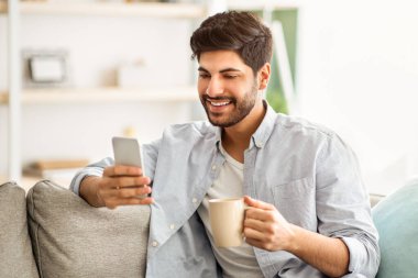 Çevrimiçi yaşam konsepti. Olumlu Arap adam elinde modern akıllı telefon ve bir fincan kahve tutarak arkadaşlarıyla sohbet ediyor, internette geziniyor ya da eğlence videosu seyrediyor.