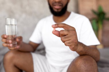 Closeup of young black man sitting on bed with glass of water, taking sleeping pills or painkillers at home. Unrecognizable millennial male using medications, holding tablet in hand, selective focus