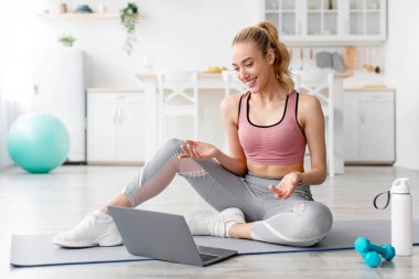 Evde çalışan koç için modern fitness aletleri. Spor giysili mutlu sarışın kadın yerde oturuyor, el kol hareketleri yapıyor ve oturma odasının iç kısmında halterler ve bir şişe su ile dizüstü bilgisayara bakıyor.