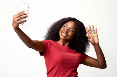 Akıllı telefonlu mutlu siyah kadın video görüşmesi yapıyor veya selfie çekiyor, beyaz stüdyo arka planının başında dikilirken Afro-Amerikan kadın el sallıyor.
