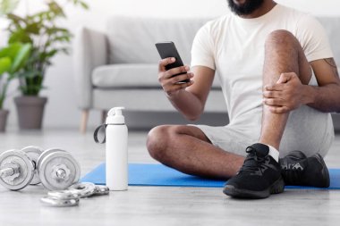 Bin yıllık Afrika kökenli Amerikalı bir adam halterlerle ve su şişesiyle halı döşemede oturuyor, akıllı telefonuyla daktilo ediyor. Spor uygulaması, spor salonu, covid-19 salgını sırasında egzersiz.