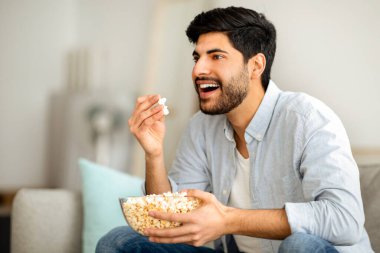 Ev eğlenceleri. Heyecanlı Arap adam patlamış mısır yiyor, televizyon izliyor, evde boş vakit geçiriyor. Oturma odasında kanepede dinlenirken sinemadan zevk alan mutlu bir adam.
