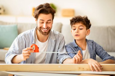 Genç babası ve küçük oğlu tahta kalaslarını bant cetveliyle ölçüyor, çocuğa ev işlerini öğretiyor, birlikte oturma odasında oturuyorlar. Ahşap işçiliği, aile hobisi ve ebeveynlik kavramı