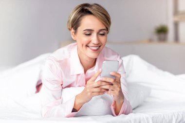 İnternette cep telefonu sörfü yapan ve evde yatakta uzanmış mesaj atan gülümseyen kadın. Akıllı telefonlu neşeli orta yaşlı kadın sosyal medyayı kolaçan ediyor. Yeni Mobil Uygulama Ters Konsepti