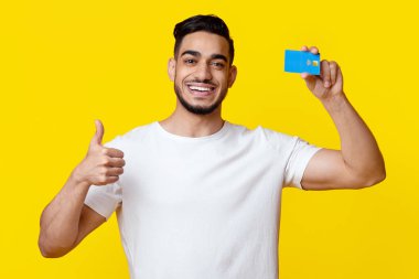 Cheerful handsome middle eastern young man holding credit card and showing thumb up, pleased client demonstrating bank product over yellow studio background. Finance, loan, savings concept