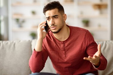 Üzgün Arap adamın kanepede oturup cep telefonuyla konuştuğu yakın plan. Sinirli ortadoğulu genç adam akıllı telefondan konuşuyor, kız arkadaşı ya da müşteri hizmetleriyle kavga ediyor.