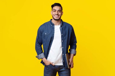 Smiling handsome middle eastern millennial man in casual outfit independent contractor posing with laptop over yellow studio background, copy space. Modern technologies concept