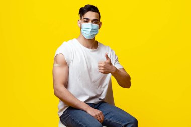 Omzunda ve başparmağında tıbbi bant olan koruyucu maskeli Arap adam Coronavirus 'a karşı aşılanmış, sarı stüdyo geçmişi, kopyalama alanı olan panorama.