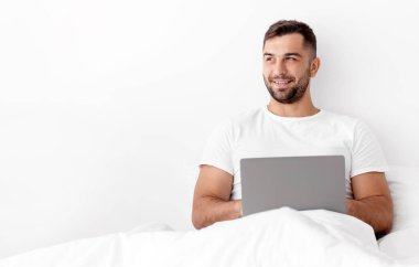 Neşeli bin yıllık beyaz kaslı yakışıklı erkek beyaz yatakta oturuyor, düşünüyor, fikir üretiyor ve bilgisayar başında beyaz arka planda yazıyor. Uzaktan sohbet, iş ve iş.