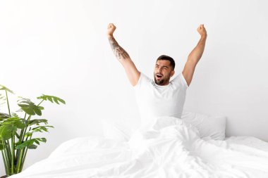 Tatmin olmuş, gülümseyen, gevşemiş, beyaz yatakta oturan, minimalist yatak odasında uzanan kaslı, kaslı, kaslı bir adam. Günaydın, iyi istirahatler ve iyi uykular, hafta sonu vadesi dolan covid-19.