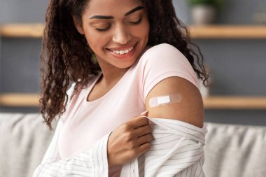 Güvenlik, koruma ve sağlık, ilaç ve bağışıklık. Neşeli bin yıllık güzel Afro-Amerikan kadın modern oturma odasındaki aşıdan sonra kollarını yara bandıyla açtı.