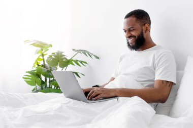 Sakallı neşeli Afro-Amerikalı erkek yatak odasındaki fotokopi odasında dizüstü bilgisayarda oturuyor. İş, video görüşmesi, iş ve iletişim çevrimiçi, kişisel izolasyon, sohbet ve yeni normal