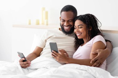 Gülümseyen Afro-Amerikan karı-koca kucaklaşmaları, yatak odasındaki beyaz yataktaki akıllı telefonlara bakmaları, boş alanlar. Sosyal medya, sohbet, yeni uygulama, covid-19 tecrit sırasında evde online teklif