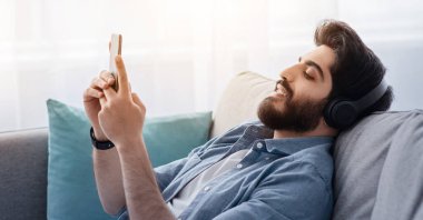 Rahat Arap adam akıllı telefondan müzik dinliyor, kablosuz kulaklıklarla iyi ses çıkarıyor, evdeki kanepede dinleniyor. Cep telefonunda müzik uygulaması kullanan sakallı adam.