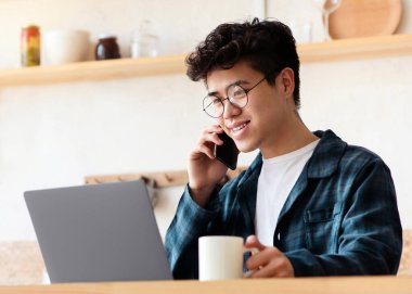 Serbest çalışma, ev ve ofisten uzaktan kumandayla karantina sırasında. Gözlüklü neşeli Asyalı genç dizüstü bilgisayarda çalışıyor, telefonla konuşuyor ve Boho tarzı mutfağında bir fincan kahve tutuyor.