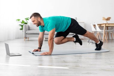 Yerde paspas üzerinde bacak egzersizi yapan ve oturma odasındaki dizüstü bilgisayara bakan mutlu, genç Avrupalı adam profili. Covid-19 salgını, spor salonu, vücut bakımı ve spor interneti, sosyal mesafe