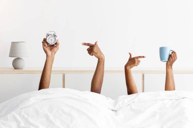 Hands of young black guy and woman stick out from under blankets, holds cups and point finger at alarm clock on bed in bedroom interior. Time to get up for work, wake up at home together, energy