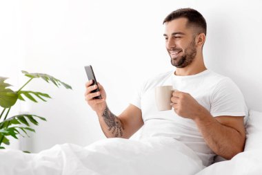 Beyaz tişörtlü mutlu beyaz beyaz beyaz adam yatak odasının içinde bir fincan kahveyle sohbet ediyor, arıyor, telefonda sörf yapıyor. Tatil ve hafta sonu, yeni uygulama, reklam ve sabah harika bir teklif.