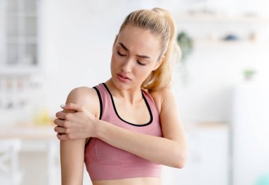 Evde antrenman, çok çalışma, antrenman ve travma sorunları. Mutsuz, bin yıllık spor kıyafetli, omuz ağrısından muzdarip, iç mekan, bulanık, kopyalama alanı olan mutsuz sarışın kadın.