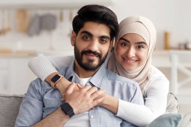 Mutlu bir aile, evde bir çift, aile fotoğrafı, online arama ve covid-19 karantina. Gülümseyen Arap kadın tesettürlü, sakallı adama sarılıyor, kanepede oturuyor, iç mekanda kopya çekiyor.